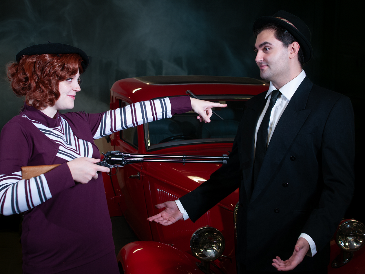 Woman with shotgun pointing at man, standing in front of red car
